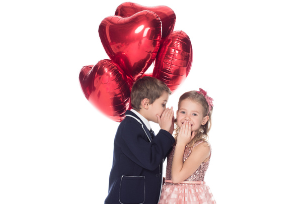 stylish little boy holding heart shaped balloons and whispering to smiling beautiful little girl isolated on white - Photo, Image