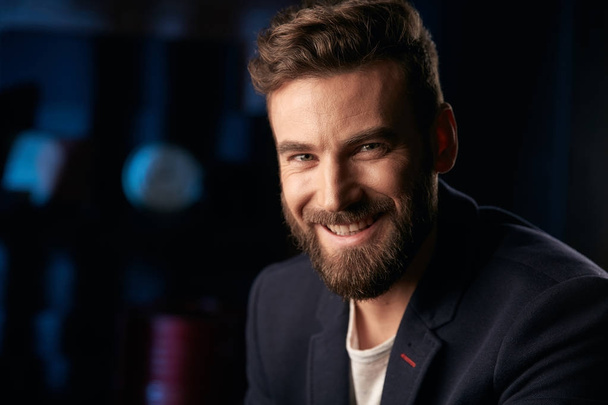 Portrait of young handsome smiling man with dark hair, beard and mustache wearing dark jacket. Studio shot with blue lighted brick background with blurred clock and red barrel - Fotoğraf, Görsel