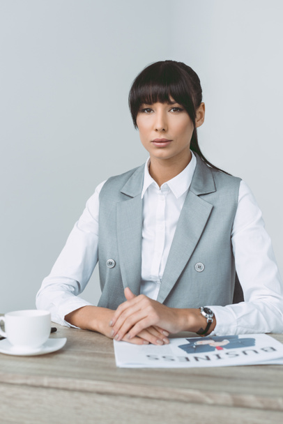 beautiful businesswoman sitting at table and looking at camera isolated on gray - Foto, afbeelding