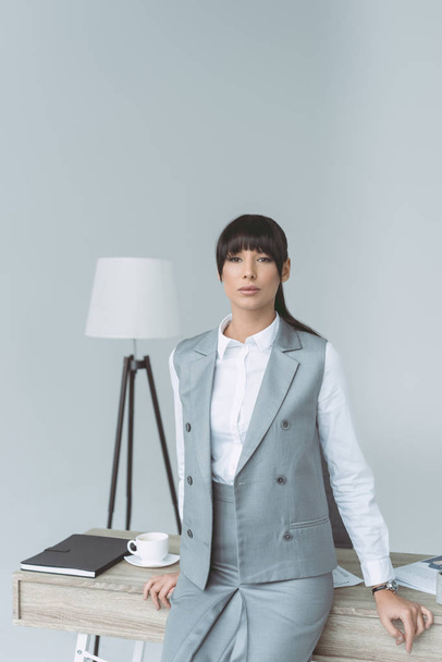 attractive businesswoman leaning on table and looking at camera isolated on gray - Photo, Image