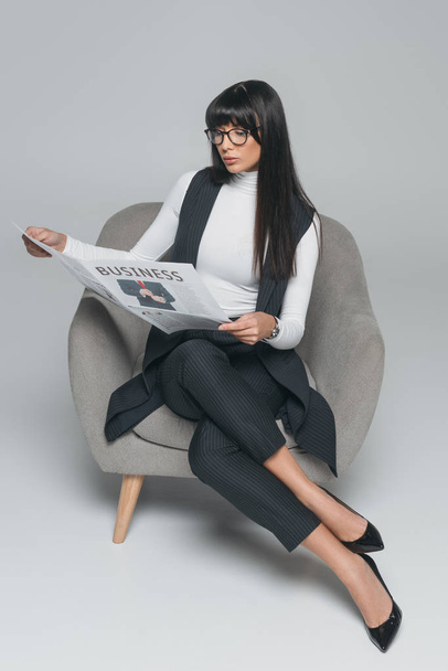 attractive brunette businesswoman reading newspaper in armchair on gray - Photo, Image