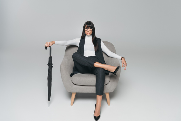 beautiful brunette businesswoman sitting on armchair with umbrella on gray - Fotoğraf, Görsel