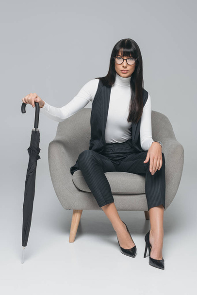 attractive brunette businesswoman sitting on armchair with umbrella on gray - Fotografie, Obrázek