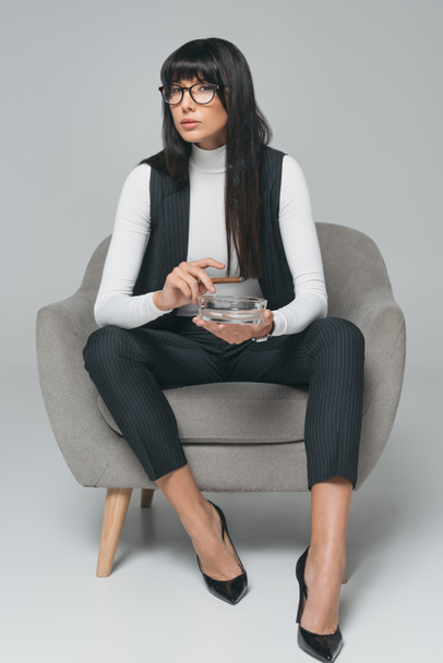 attractive brunette businesswoman holding ashtray and cigar on gray - Valokuva, kuva