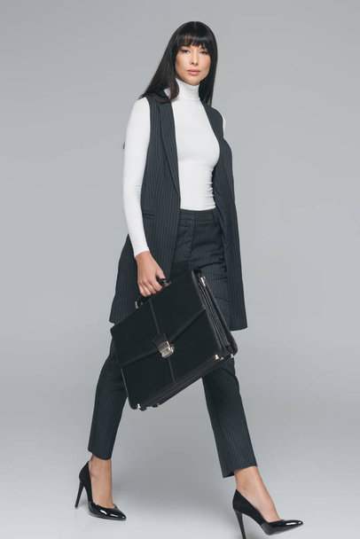 attractive brunette businesswoman walking with briefcase on gray - Fotografie, Obrázek