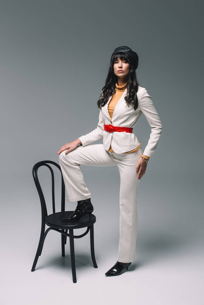 beautiful brunette woman in white suit putting leg on chair on gray - Photo, Image