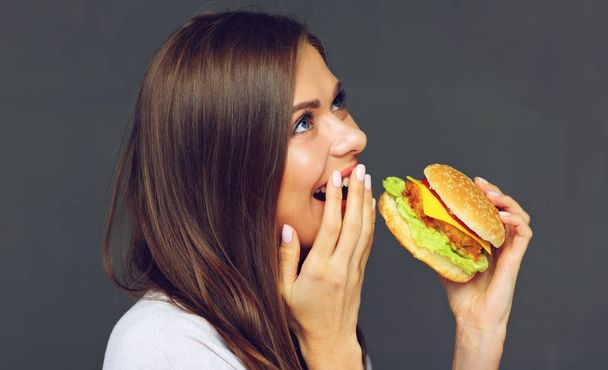 Young woman eating burger fast food. Isolated portrait against gray wall. - Zdjęcie, obraz