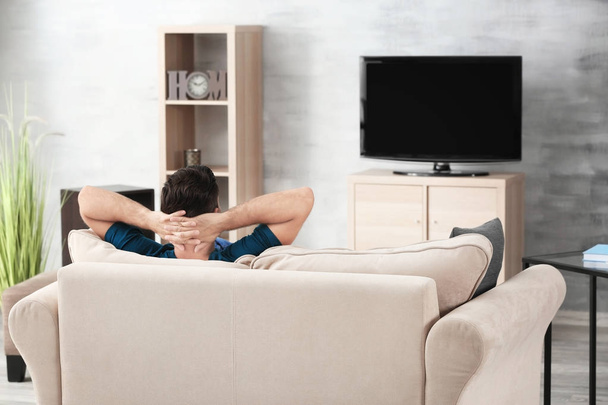 Красивый мужчина смотрит телевизор, отдыхая дома на диване
 - Фото, изображение
