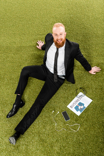 high angle view of bearded businessman smiling at camera while sitting on green grass with smartphone, earphones, eyeglasses and newspaper  - Fotó, kép