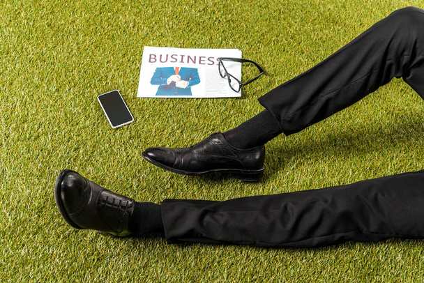 částečný pohled mužských nohou ve stylové kožené boty, brýle, noviny a smartphone s prázdnou obrazovkou na zelené trávě - Fotografie, Obrázek