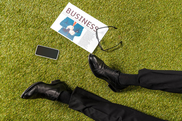 обрізаний знімок чоловічих ніг у стильному шкіряному взутті, окулярах, газеті та смартфоні з порожнім екраном на зеленій траві
 - Фото, зображення