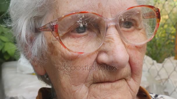 Vanha nainen silmälaseissa katsoo eteenpäin ja hymyilee. Muotokuva onnellinen mummo silmälasit viettää aikaa ulkona. Isoäidin tunteet. Sulje hitausliike - Materiaali, video