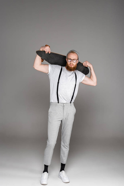 handsome stylish bearded man holding longboard on shoulders and looking at camera on grey  - Φωτογραφία, εικόνα