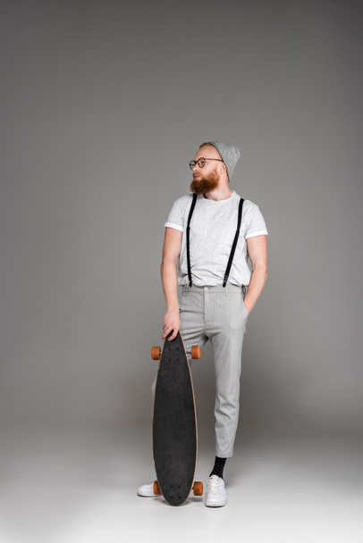 handsome stylish bearded man standing with skateboard and looking away on grey - Zdjęcie, obraz
