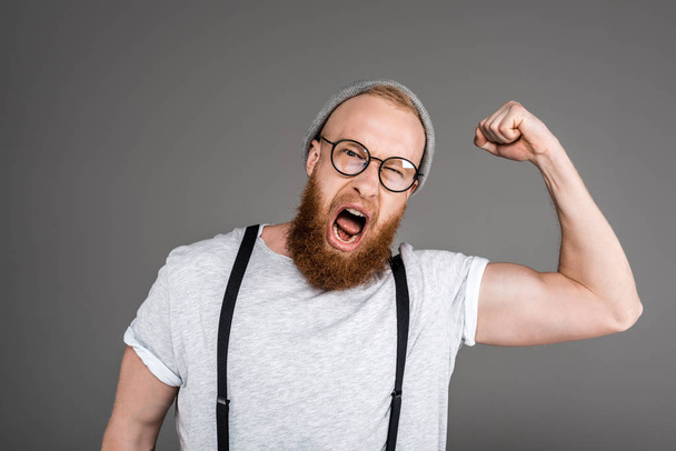 excited bearded man showing biceps and screaming at camera isolated on grey - Fotó, kép