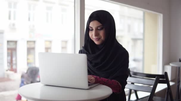 Attractive woman in hijab searching for something in internet. Portrait of smiling young muslim woman working on modern laptop in cafe. Studying or working. Slowmotion shot - Video, Çekim
