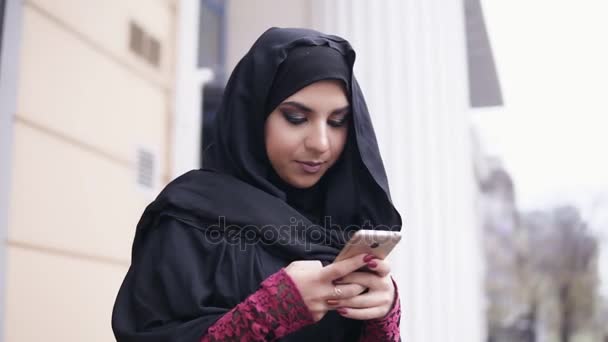 Camera round movement: Young attractive woman wearing hijab standing in the street, typing a message on her mobile phone. Slowmotion shot - Materiał filmowy, wideo