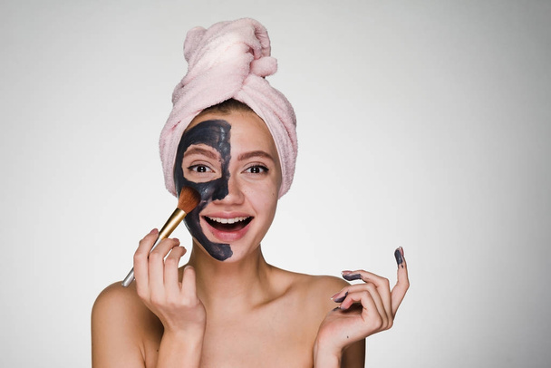 Laughing beautiful girl with a pink towel on her head puts on half face clay mask - Fotoğraf, Görsel
