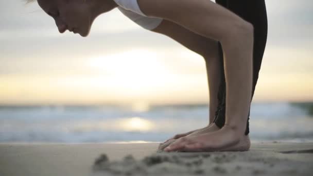 Mujer de yoga flexible que se estira hacia adelante dobla tocando la frente hasta las rodillas al aire libre al atardecer playa de mar de cerca. Instructor de fitness gimnasta profesional realiza asana Uttanasana de pie descalzo sobre la arena
 - Metraje, vídeo