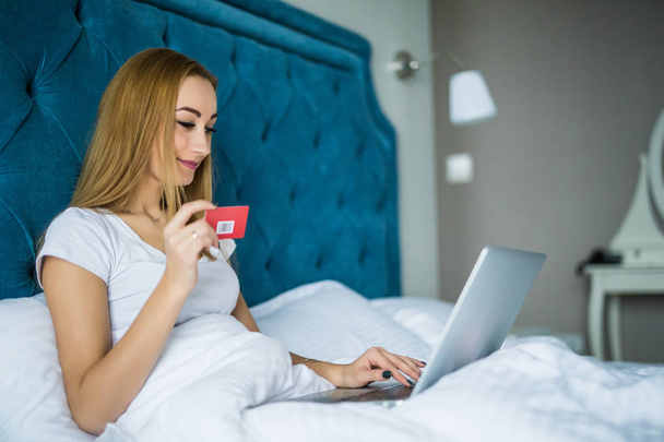 Smiling woman lying on her bed with her credit card in hand as she uses her laptop while wake up in the morning - Fotoğraf, Görsel