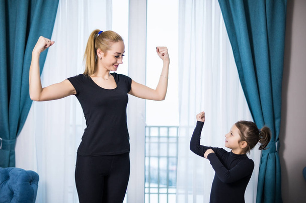 Hermosa joven y encantadora hija pequeña están mostrando sus bíceps y sonriendo mientras hacen ejercicio en casa
 - Foto, Imagen