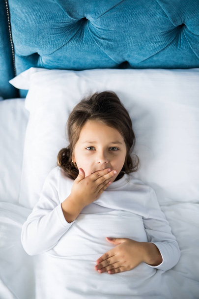 Vista superior de la niña bosteza con los ojos del armario, cubriéndose la boca mientras está acostada en la cama en casa
 - Foto, Imagen