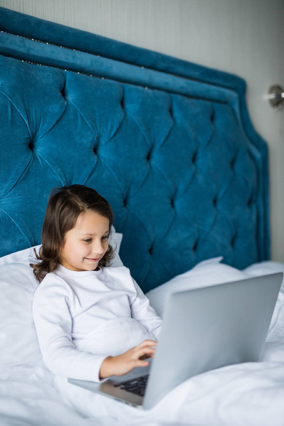 Spokojený nadšené malá holčička seděla na posteli a usmívala se na kameru, zatímco notebooku umístěny před ní - Fotografie, Obrázek