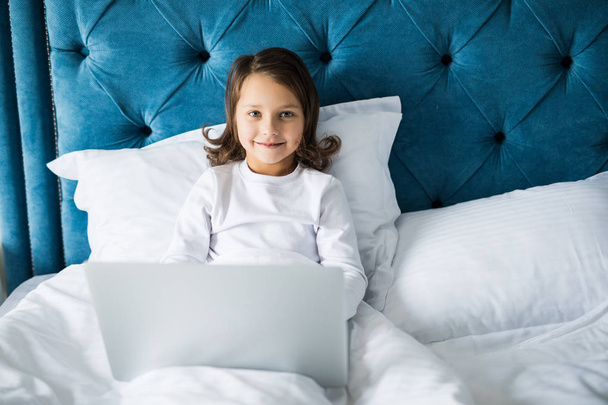 Zadowolony entuzjastycznie dziewczynka siedzi na łóżku podczas uśmiechając się do kamery, podczas gdy laptop umieszczone przed nią - Zdjęcie, obraz