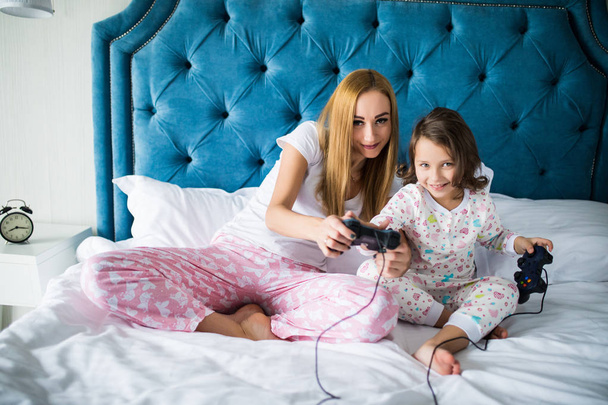 Cheerful mother and daughter playing video games in bed at home - 写真・画像