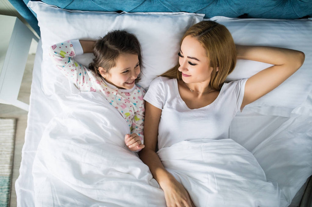 Vista superior de una mujer sonriente feliz divirtiéndose con su hija mientras están acostados juntos en la cama
 - Foto, Imagen