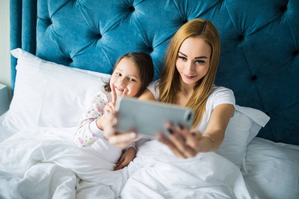 Anne ve kızı yatakta yatarken ve smartphone ile kendi kendine portre çekici. Yatak Odası, küçük bir kız ile selfie alarak kadın. - Fotoğraf, Görsel
