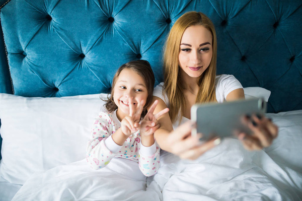 Madre e hija tumbadas en la cama y tomando autorretrato con smartphone. Mujer tomando selfie con una niña en el dormitorio
. - Foto, imagen