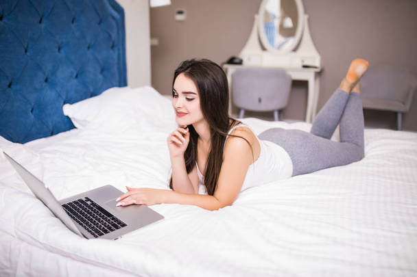 Mujer bonita sonriente poniéndose al día en sus redes sociales mientras se relaja en la cama con una computadora portátil en un día perezoso
 - Foto, imagen