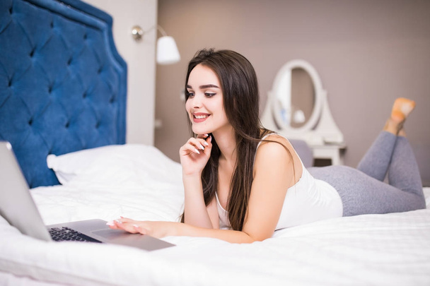 Dość uśmiechający się kobieta zajęty, przeglądania na laptopie przy filiżance kawy na białe łóżko. - Zdjęcie, obraz