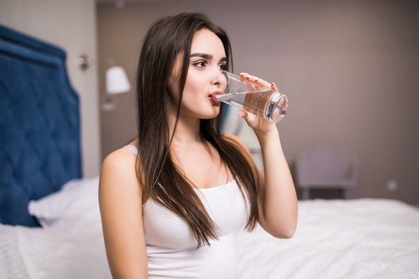 Šťastná mladá žena drží sklenici vody při sezení na posteli doma - Fotografie, Obrázek