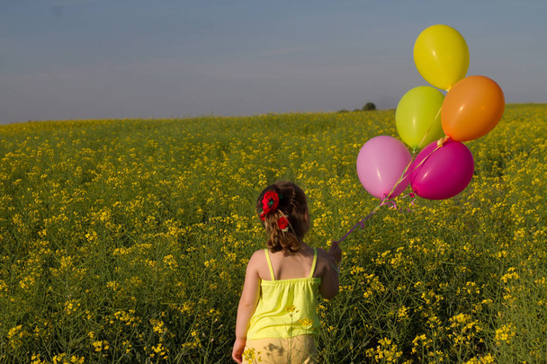 kleines Mädchen in niedlichem Kleid, das bei Sonnenuntergang im goldenen Weizenfeld läuft und Luftballons in der Hand hält. Blick von hinten. - Foto, Bild