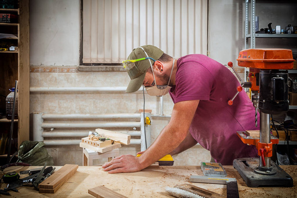 Mladý silný muž stavitel tesař pracuje s dřevěnou tyčí pro výrobu nábytku, opatření a řezů v dílně, v pozadí mnoha nástrojů a pracovních podmínek - Fotografie, Obrázek
