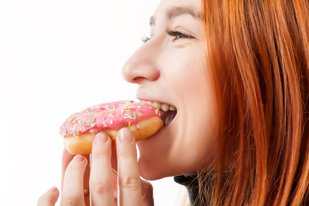 Mladá zrzavá žena v Černý rolák a Kuchyňské zástěry se směje a jí donut růžový s radostí na bílém pozadí izolované - Fotografie, Obrázek