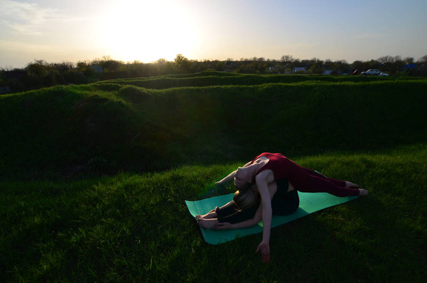 Dos jóvenes de pelo rubio en trajes deportivos practican yoga en una pintoresca colina verde al aire libre por la noche. El concepto de ejercicio deportivo al aire libre
 - Foto, imagen