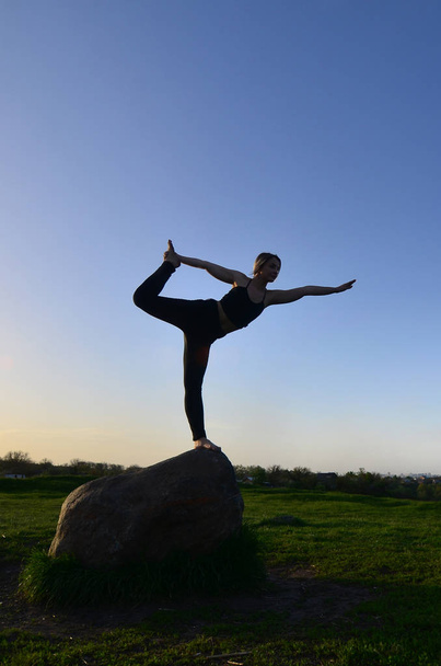 Silueta de joven rubia en traje deportivo practica yoga en una pintoresca colina verde por la noche al atardecer. El concepto de ejercicio y estilos de vida saludables
 - Foto, Imagen
