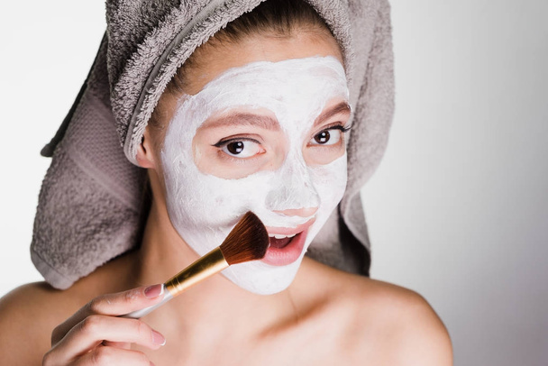 lovely beautiful girl with a towel applied a white nutritious mask to her face with a brush - Photo, Image