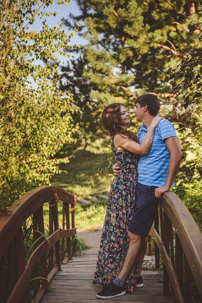 Happy romantic couple in the village, stroll on the wooden bridge. Young beautiful woman in long summer dress hugging the man. - Valokuva, kuva
