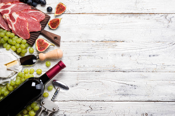 Red wine, grape, figs, cheese camembert, prosciutto meat on white wooden background. Appetizer, gourmet, romantic concept - Fotó, kép