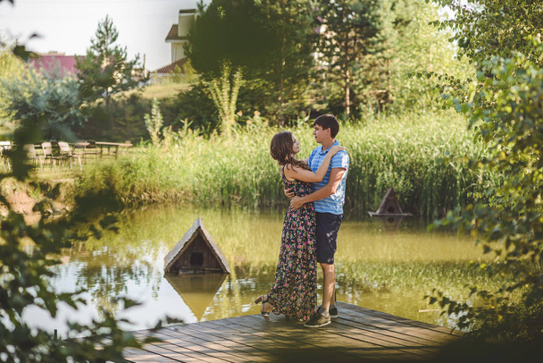 Happy romantic couple in the village, stroll on the wooden bridge near the lake. Young beautiful woman and man hugging each other. - Photo, Image