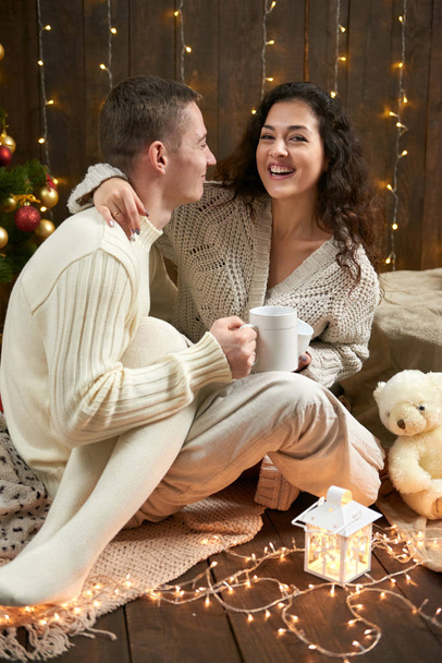 Happy couple drinking tea and talking in christmas decoration, sitting on floor in dark wooden interior with lights. Romantic evening and love concept. New year holiday. - Photo, Image