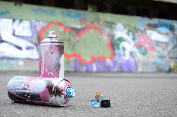 Mehrere gebrauchte Spraydosen mit rosa und weißer Farbe und Kappen zum Sprühen von Farbe unter Druck liegen in farbigen Graffiti-Zeichnungen auf dem Asphalt neben der bemalten Wand. - Foto, Bild