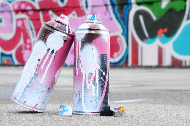 Diverse gebruikte spuitbussen met roze en witte verf en caps voor het spuiten van verf onder druk ligt op het asfalt in de buurt van de geschilderde muur in gekleurde graffiti tekeningen - Foto, afbeelding