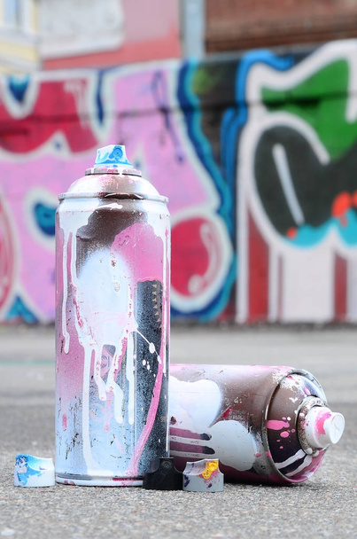Varias latas de spray usadas con pintura rosa y blanca y tapas para rociar pintura bajo presión se encuentra en el asfalto cerca de la pared pintada en dibujos de graffiti de colores
 - Foto, Imagen