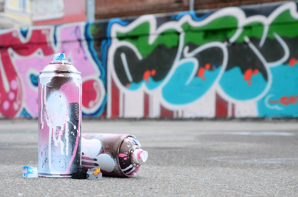 Plusieurs pulvérisateurs utilisés avec de la peinture rose et blanche et des capuchons pour pulvériser de la peinture sous pression se trouve sur l'asphalte près du mur peint dans des dessins de graffiti colorés
 - Photo, image