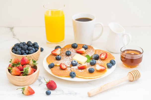 gesundes Frühstücksset mit Pfannkuchen aus Blaubeeren und Erdbeeren und schwarzem Kaffee, Milch und Orangensaft auf weißem Steintischhintergrund - Foto, Bild
